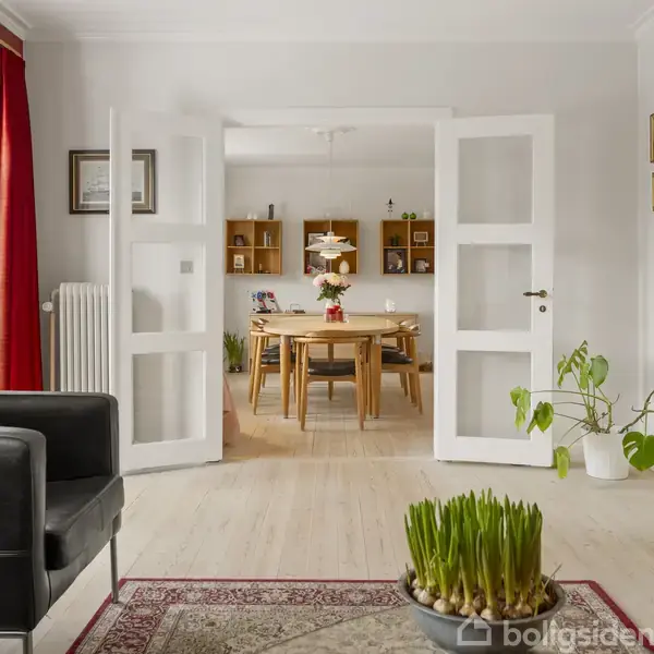 Dobbelt glasdøre åbner mod spisestue med træbord og stole. Rummet har lyse vægge, gulvtæppe, potteplanter, røde gardiner og væghængte hylder. Lysekrone hænger over bordet.