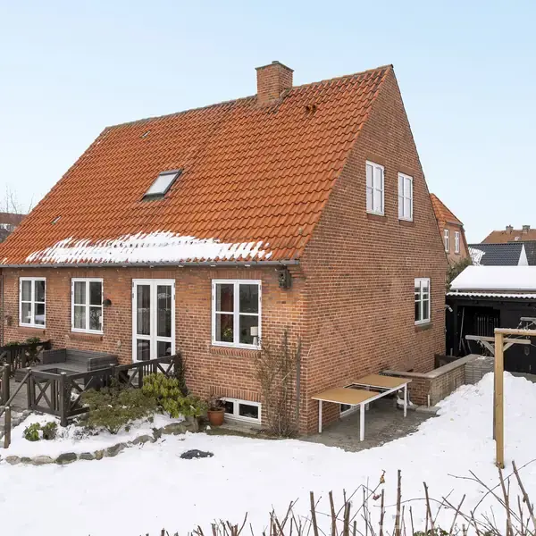 Et rødt murstenshus med rødt tegltag står i et snedækket landskab. Foran huset er en lille have med et træhegn. En carport findes på husets højre side.