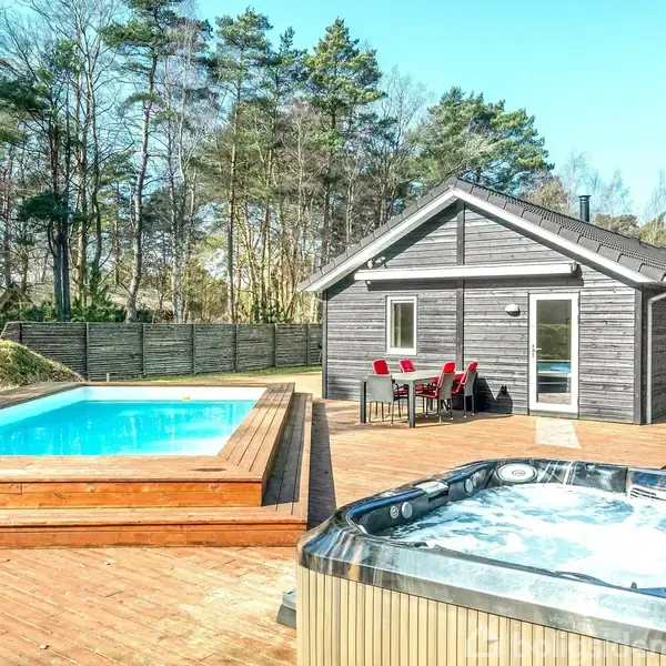 Træhus står i skovklædt have med udendørs pool og boblebad på træterrasse. Røde stole og bord ses ved indgangen, skov omgiver området.