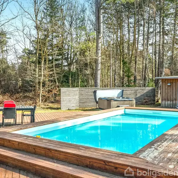 En rektangulær swimmingpool står fyldt med blåt vand på en terrasse af træ. Omkring poolen er der skov og et mørkt træhus med røde og sorte borde og stole.