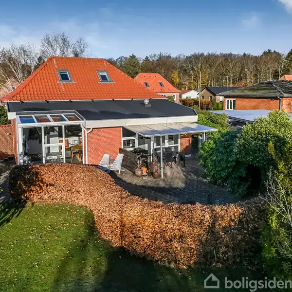 Hus med rødt tag står stille i en grøn have, omgivet af buske. En terrasse med glasoverdækning er synlig, og der er havemøbler. Baggrunden viser flere huse og skov.