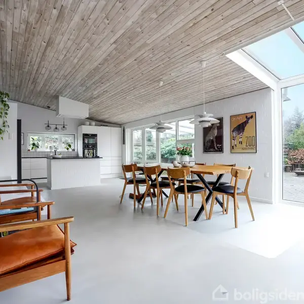 Stue med spisebord og stole placeret på lyst gulv; køkken i baggrunden og store vinduer mod højre giver udsigt til vegetation udenfor. To lænestole står ved væggen til venstre.