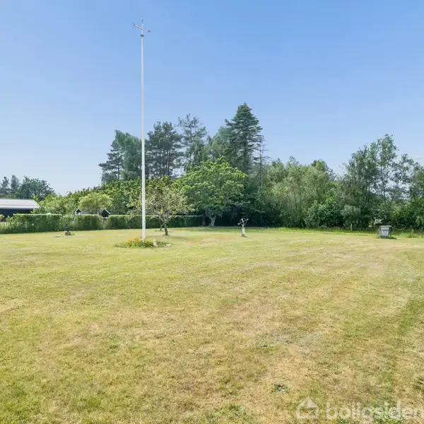 Flagstang står frit på en stor, åben græsplæne; omkranses af tæt bevoksning og træer. Til højre ses en del af et murstenshus med vinduer og en parabolantenne.