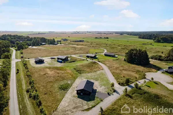 Et sort hus ligger i forgrunden, omgivet af grusveje og grønne marker med flere huse spredt ud i et landligt landskab under en blå himmel.