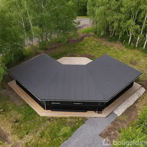 Sort, moderne hus med fladt tag er placeret på græsareal omgivet af træer. Huset har en vinkelformet struktur og er omkranset af en terrasse. Stier fører til vejen.