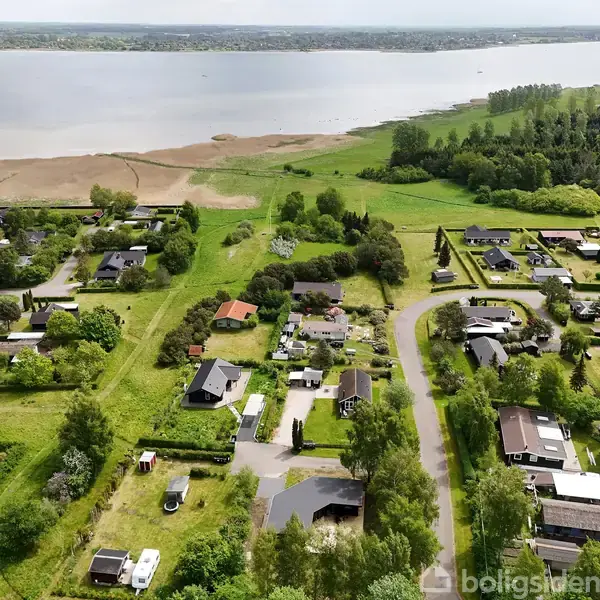 Et boligområde med spredte huse omgivet af grønne græsplæner og træer. Veje forbinder husene, og i baggrunden ses en bred søbred med vandet strækkende sig til horisonten.