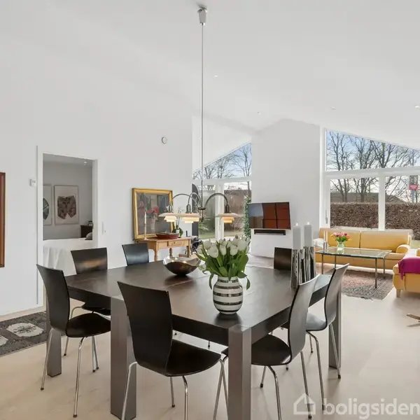 Et sort spisebord med otte stole står centralt i et lyst rum. På bordet er blomster og lysdekorationer. Store vinduer giver udsigt til en træbevokset udendørs have.
