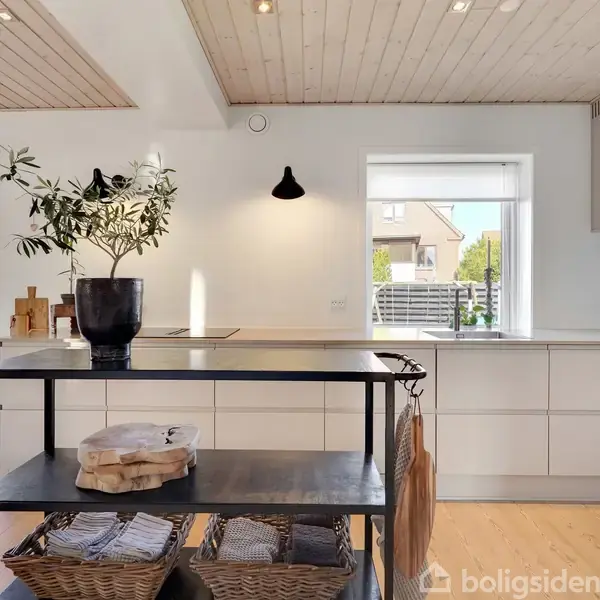 Et moderne køkken med en sort reol, der har kurvefyld og en potteplante ovenpå. Reolen står mellem hvidt køkkenskab og spisebord i et lyst rum med trægulv.