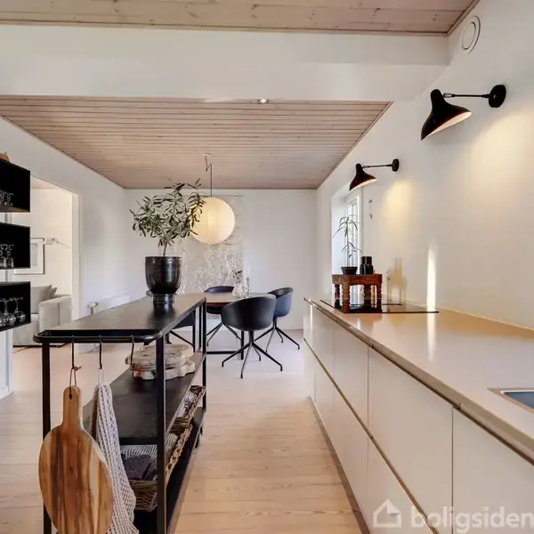 Et lyst køkken med en lang bordplade og indbygget vask. En ø med planter og opbevaring står i midten. Hylder med glas ses i baggrunden.