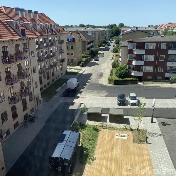 En hvid varevogn står parkeret i et boligområde med flere lejlighedsbygninger omkring. Der er cykler op ad væggen, og mindre biler på vejen. Det er solrigt.