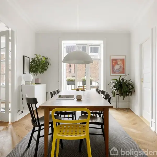 Et spisebord med sorte og gule stole står i et lyst spisestueområde. En stor lampe hænger over bordet, og der er planter og dekorationer omkring. En dør åbner til stuen.