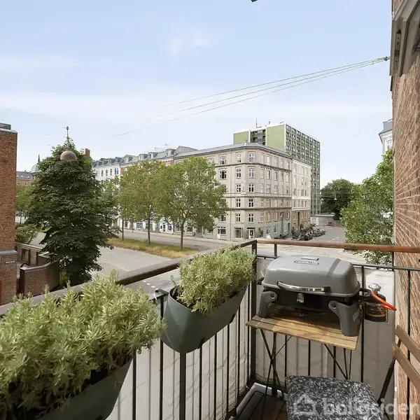Balkon med en grill på et bord, omkranset af planter i blomsterkasser. Baggrunden viser en byvej med grønne træer og historiske bygninger under en klar himmel.