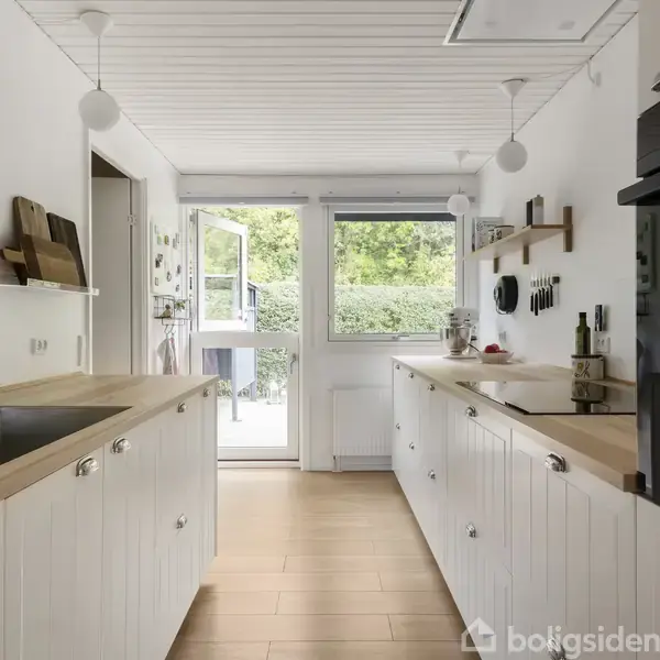 Køkken med hvide skabe og træbordplader. Vasken til venstre, ovn til højre. Åben dør mod terrasse og have i baggrunden. Hængende lys og væghylder.