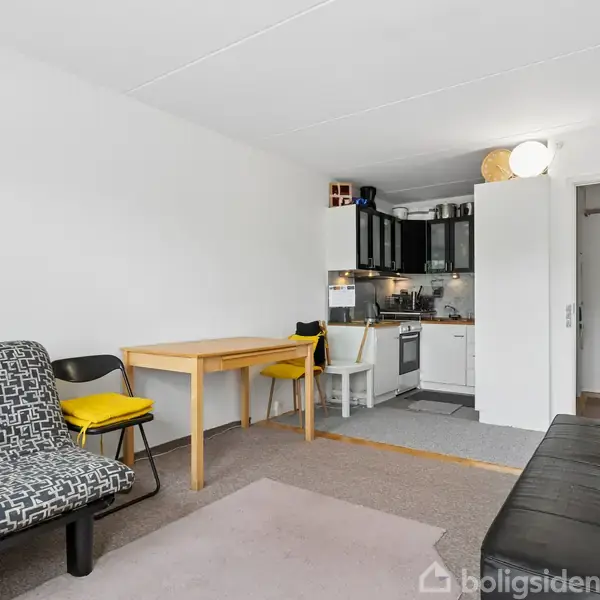 Sort-hvid sovesofa står i et lyst stueområde med trægulv. Spisebord med to gule stole er til højre, overfor et lille køkken med hvide skabe.