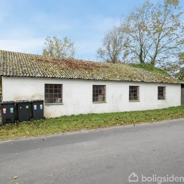 En hvid bygning med fladt tag står stille ved en vej, omgivet af træer. Tre affaldscontainere er placeret til venstre for bygningen.