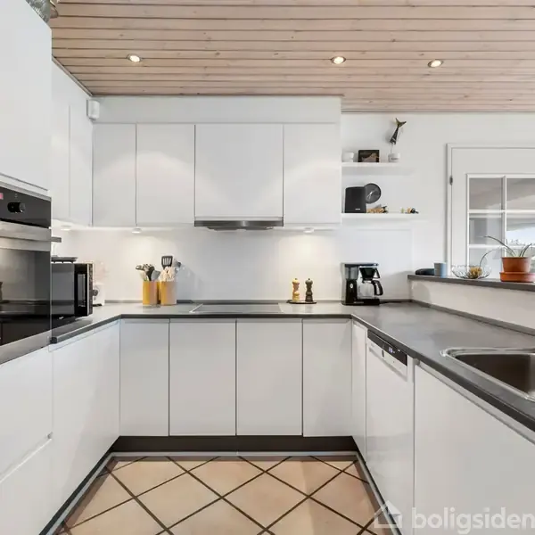Køkken med hvide skabe og sorte bordplader, indbygget ovn og vask, pænt oplyst af spotlys i loftet af træ. Der står planter og køkkenudstyr på bordpladen.