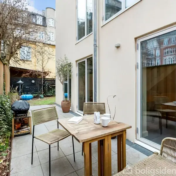 Et bord med kaffekopper og stole står i en smal baggård. Der er en grill og planter langs et træhegn, med moderne bygninger i baggrunden under en blå himmel.