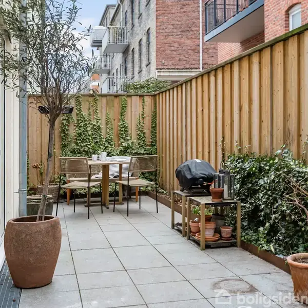 Terrasse med flisebelægning, bord og stole i træ i midten, omgivet af grønne planter. Til højre står en grill og flere urtepotter. Omgivet af høje hegn og bygninger.