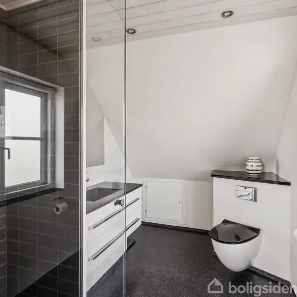 Et minimalistisk badeværelse med en bruseniche til venstre og et toilet til højre. Badeværelset har en skråvæg, sort gulv, og hvide skabe under en sort bordplade.