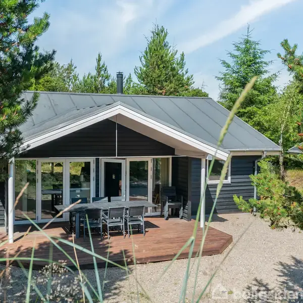 Et sommerhus med sort træbeklædning og grått tag står stille omgivet af grønne træer. På træterrassen foran huset er der et spisebord med stole.