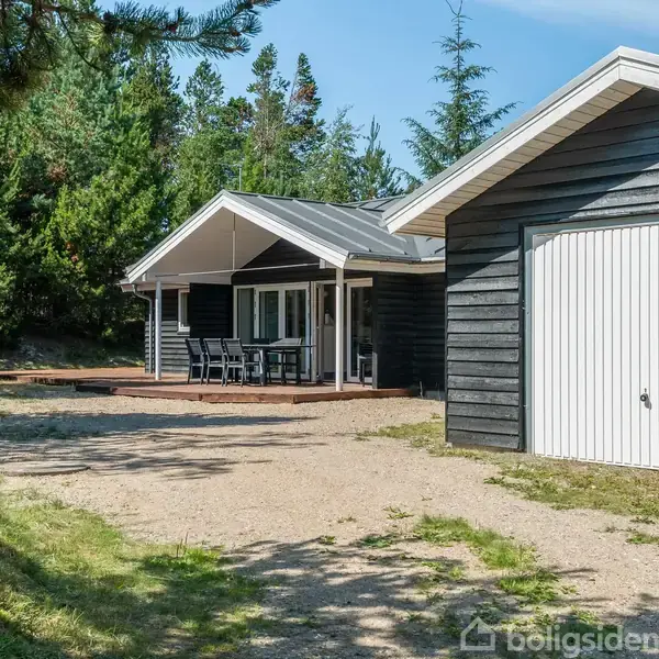 Sommerhus med træbeklædning står omgivet af skov. En overdækket terrasse foran huset har et bord og stole. Til højre er en garage med en hvid port.