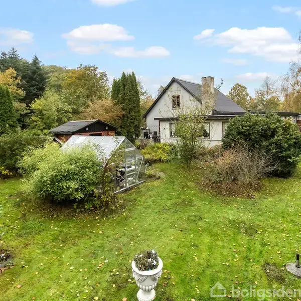Et hvidt hus med sort tag står omringet af frodig have og grønne planter. Et drivhus ses midt i haven. Blå himmel med skyer over skovklædte områder.