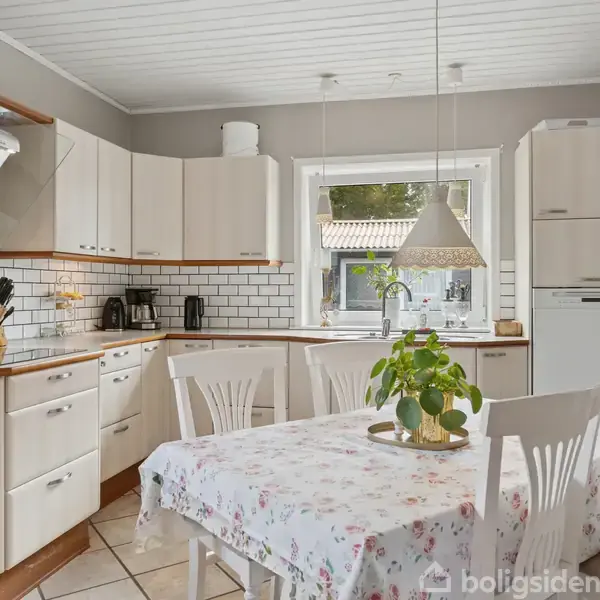 Køkkenbord med blomstret dug og hvidmalede stole står i centrum, omgivet af hvide køkkenskabe. Et vindue med udsigt til haven slipper lys ind. Køkkenet har flisebelagt væg og rustfri emhætte.