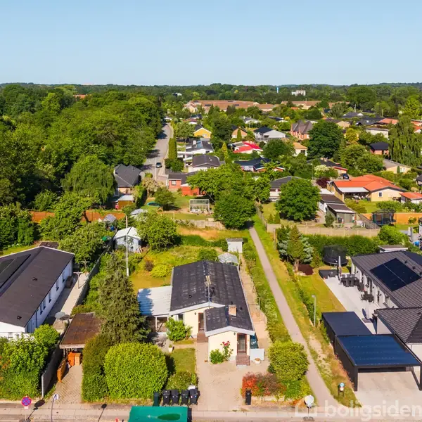 Boligkvarter med huse omgivet af grønne haver. Huse i forskellige størrelser og tags farver ses fra luften, med veje og træer i baggrunden.