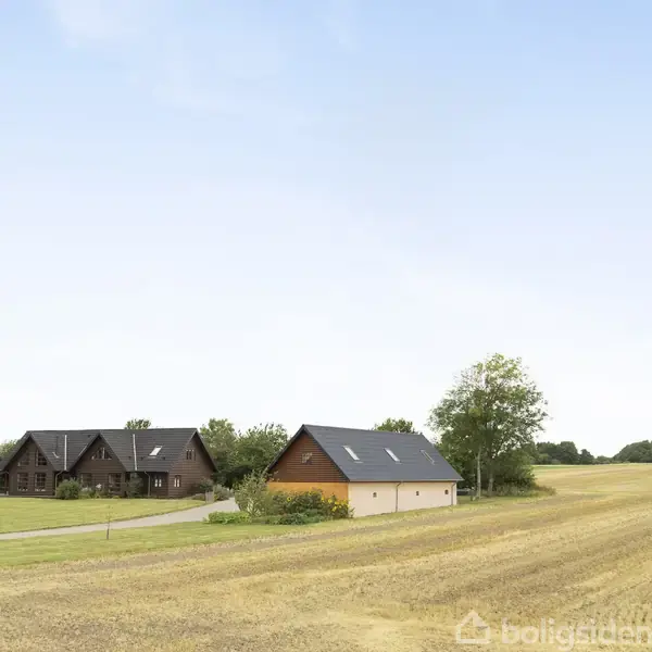To huse står ved siden af hinanden på en grøn grund ved siden af en høstet mark. En blå himmel ses i baggrunden.