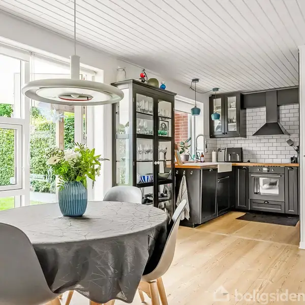 Rundt spisebord med vase og blomster står i lys spisestue med vinduer til have. Køkken med sorte skabe og hvid flisevæg ses i baggrunden.