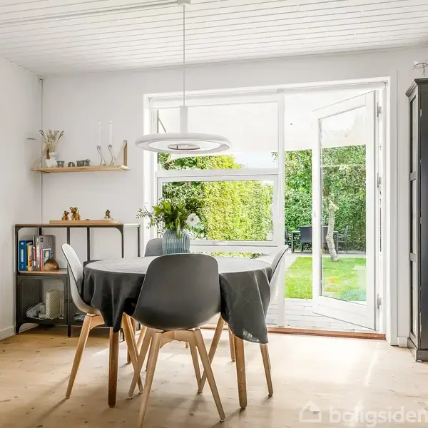 Et spisebord med fire stole er omgivet af hylder og et skab med glas. I baggrunden er der en åben glasdør, der fører ud til en have med græsplæne.