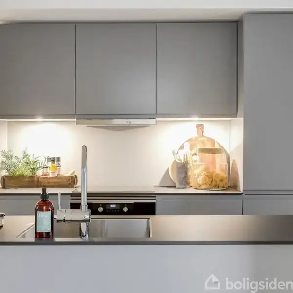 Gråt moderne køkken med skabsfronter. Bordpladen har en vask og diverse flasker. Belysning under skabe. Dekorative elementer som en plante og blomster i vasen skaber hygge.