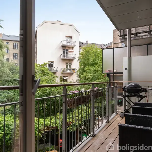 En balkon med trægulv har to sorte møbler og en grill. Omgivet af grønne træer og udsigt til nabobygninger i en bymæssig baggård.