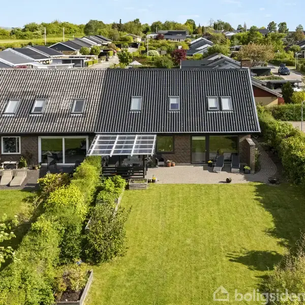 Rækkehus med sort tag ligger i et villakvarter, omgivet af velplejede haver og grønne marker. Foran huset er der en stor, græsplæne med terrasse og solbeskinnede områder.