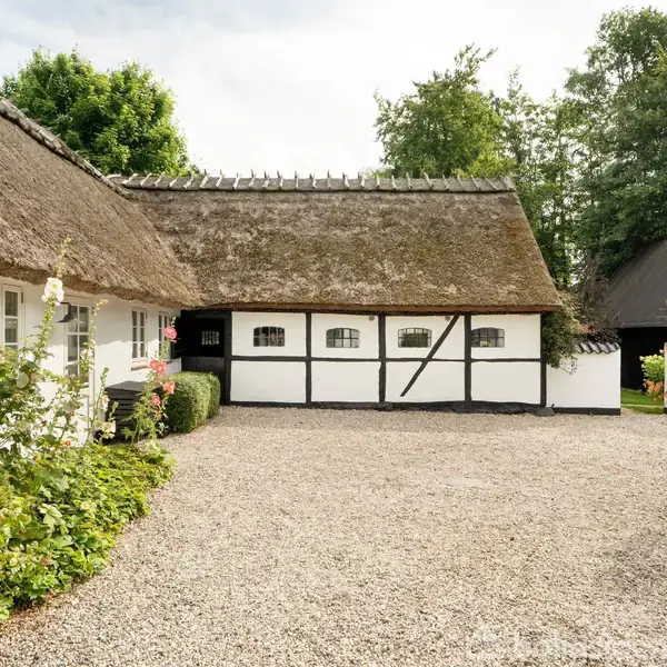 Stråtækt hus omgivet af grusgårdsplads, med hvidkalkede vægge og sort bindingsværk. Grønne planter og træer ses i baggrunden, hvilket skaber en idyllisk, landlig atmosfære.