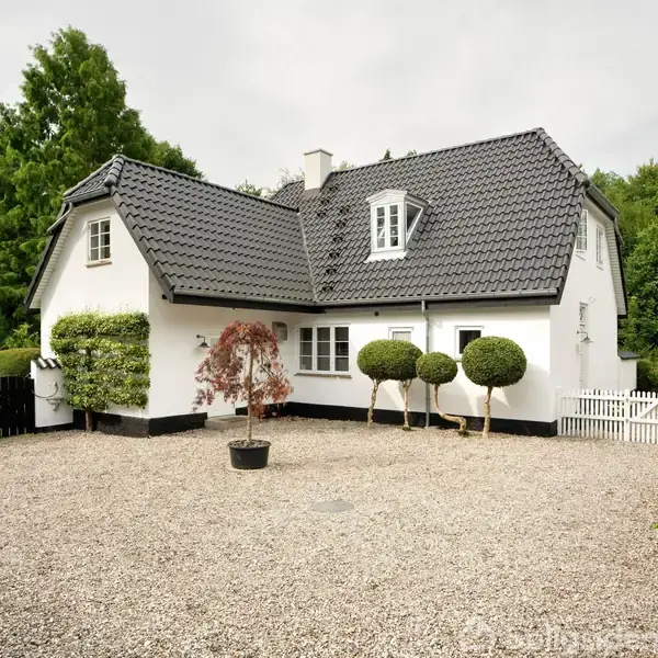 Et hvidt hus med sort tag står i en grusbelagt gårdsplads, omgivet af grønne træer og buske. Huset har små vinduer, og pladsen har en dekorativ plante i midten.