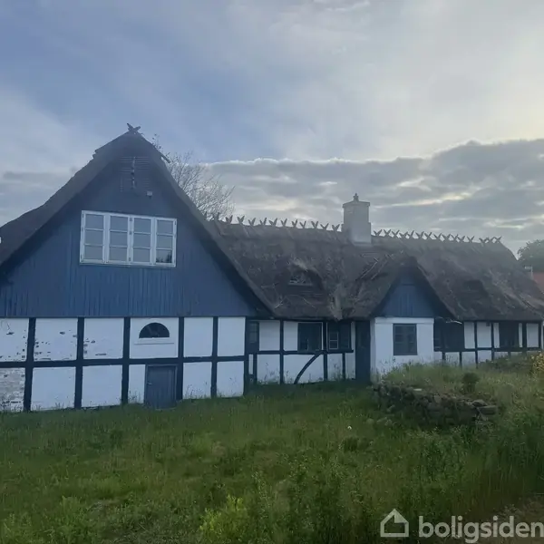 Bindingsværkshus står stille i en frodig have med græs, har stråtag og blå-hvide vægge. Skorstensrør stikker ud fra taget under en overskyet himmel.