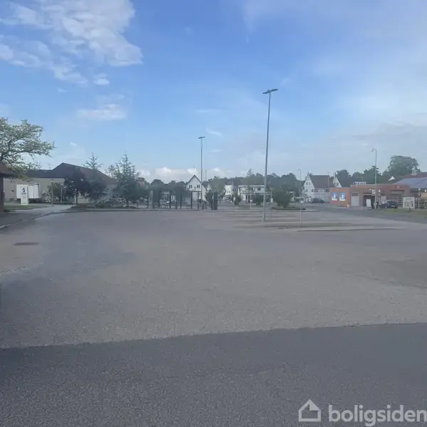 En bil holder parkeret i et næsten tomt, asfalteret område ved siden af en række bygninger. Baggrunden viser et par træer under en blå himmel med lette skyer.