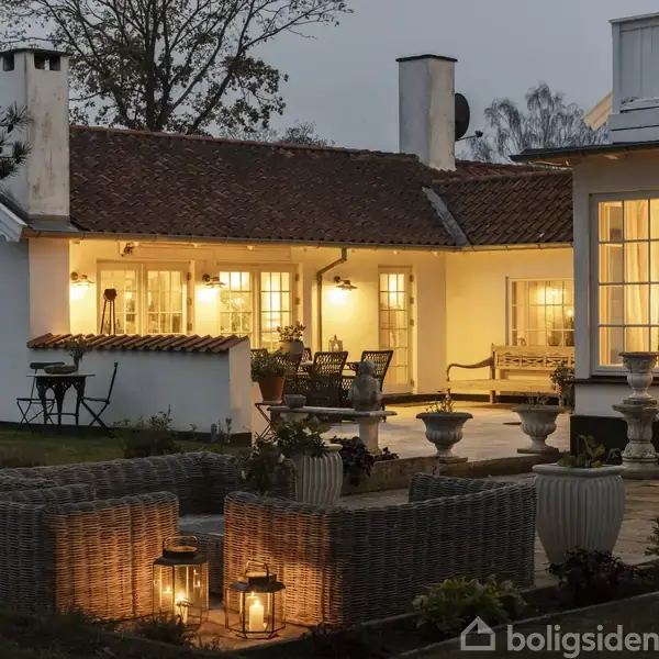Et hyggeligt hus med oplyst terrasse, omgivet af kurvestole og lanterner. Haven har plantekasser, og omgivelserne er rolige med træer i baggrunden.