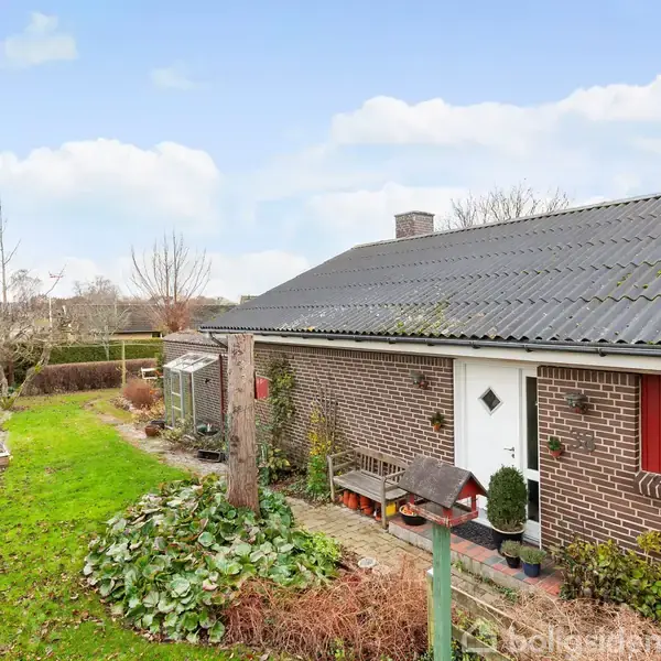 En lav, murstensbolig med sort tag ligger ved en velplejet have med græsplæne og buske. Ved siden af huset ses en lille sti og et fuglehus.