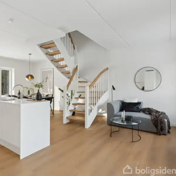 En moderne stue med en hvid trappe, der fører opad. En sofa med puder står til højre, og der er et rundt spejl på væggen. Rummet har store vinduer med udsigt.