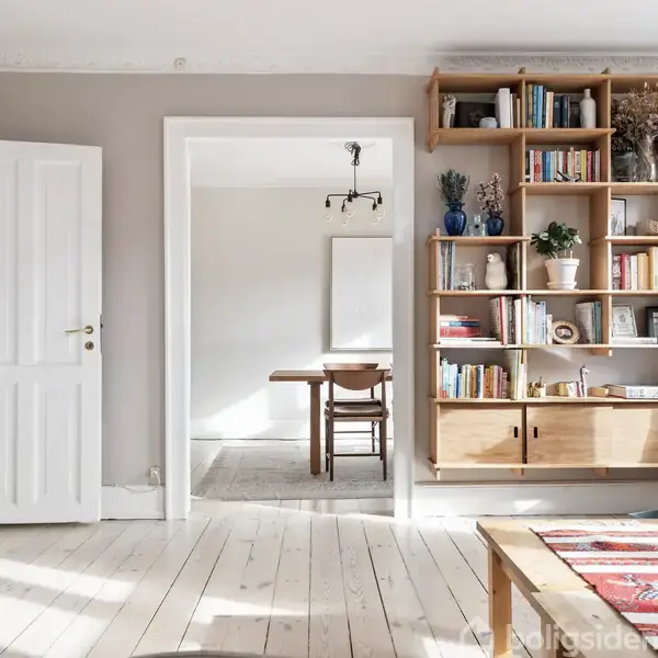 Træreol fyldt med bøger og planter står i stuen, ved siden af en åben dør, der fører til et lyst spisestueområde med et bord og en stol.