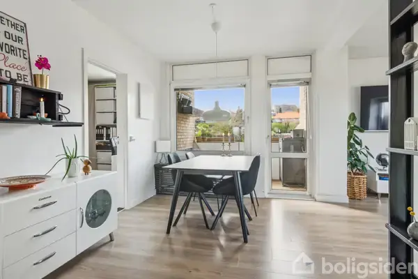 Spisebord med fire stole står i lys spisestue, hvor en dør fører til en altan. En hvid skænk med dekorationer står til venstre. Ingen tekst på billedet.