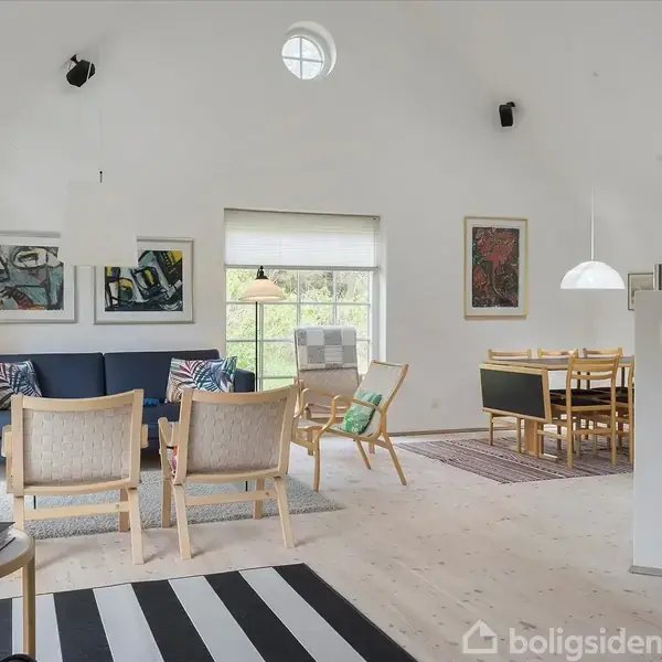 Stue med blå sofa og træstole, placeret foran et stort vindue med udsigt. Væggene er dekoreret med kunst, og der ses et spisebord i baggrunden. Gulvet er lyst trægulv.