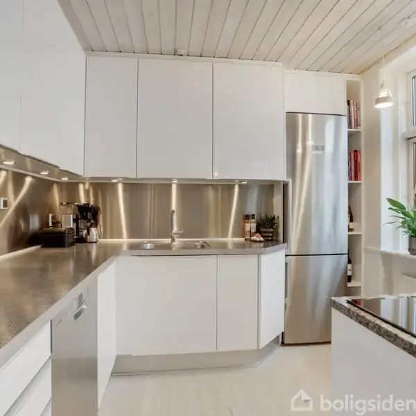 Køkken med hvide skabe og stålbordplader, stilfuldt oplyst af indbyggede lys, med en indbygget ovn og et køleskab ved vinduet. Udsigt til murstensbygninger udenfor.