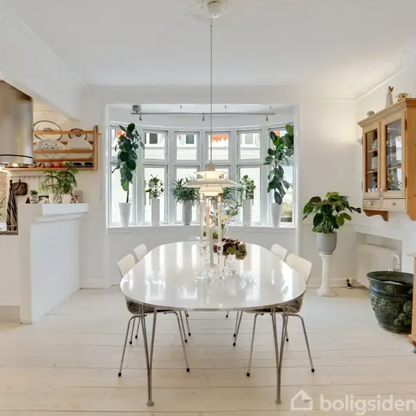 Et spisebord med seks stole står i et lyst rum, omgivet af grønne planter og hvide vægge. Der er et stort vindue i baggrunden, der lukker naturligt lys ind.