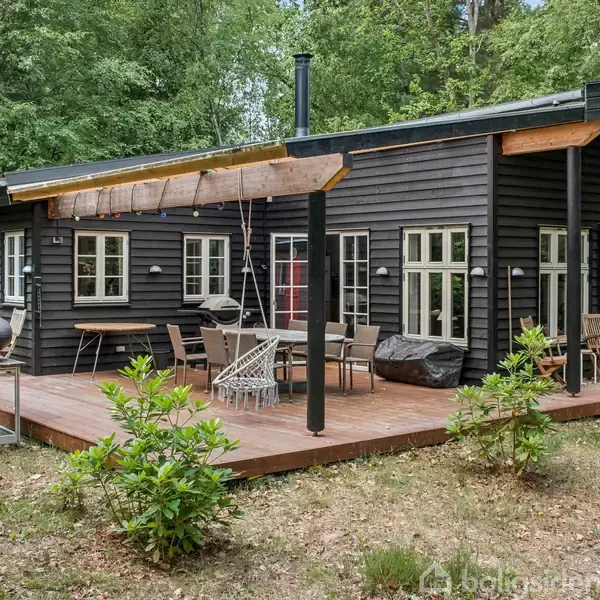 Sort træhus med store vinduer, står midt i grønt skovområde. På terrassen er der haveborde, stole og en grill, omgivet af træer og buske.