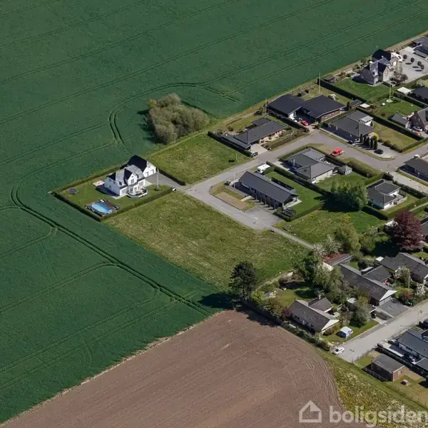 Et hvidt hus med en swimmingpool står isoleret ved siden af en stor mark. En mindre boligkvarter med huse og veje ligger til højre for huset.