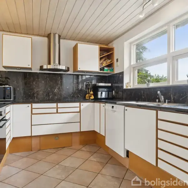 Et moderne køkken med hvide skabe og sort marmorbordplade. Et vindue lader lys ind. Køkkenet indeholder en mikroovn, kaffemaskine og planter, der bidrager til en lys og indbydende atmosfære.