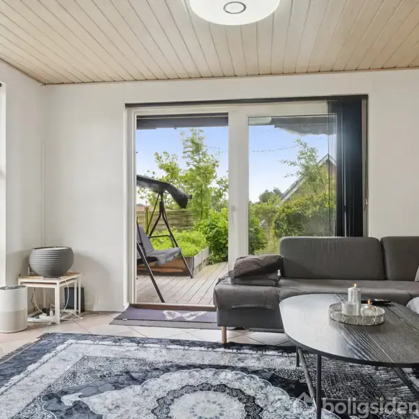 Grå hjørnesofa står i en lys stue mod en havedør. Udenfor ses en terrasse med en hængestol. Rummet er indrettet med grønne planter og et dekorativt tæppe.